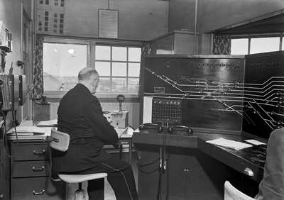 159672 Interieur van het seinhuis van de N.S. te 's-Hertogenbosch met de treindienstleider achter het NX-bedieningstableau.